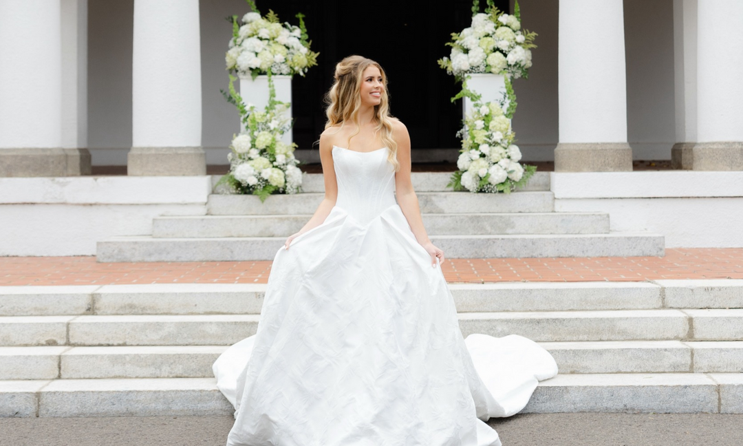 bride in white strapless wedding dress with basque waist and jacquard detailing, outside in front of steps into venue and white floral arrangments 