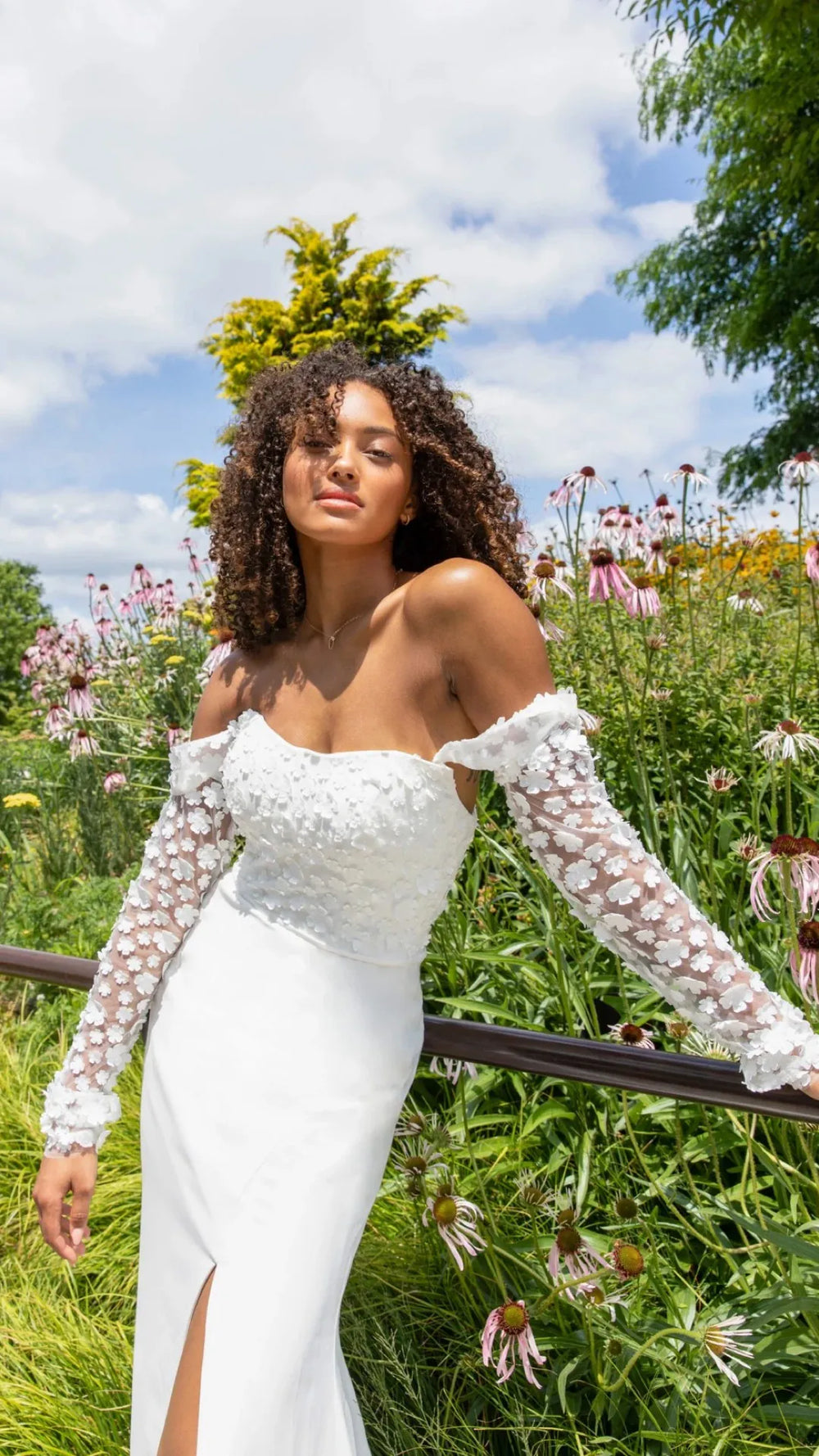 A woman wearing a white off-the-shoulder wedding dress with long sheer lace sleeves, standing outdoors in a flower garden under a blue sky.