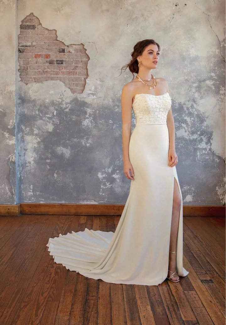 A woman modeling a strapless ivory wedding gown with a fitted silhouette, lace bodice, high side slit, and long train, standing in a room with a distressed gray wall and wood floor.