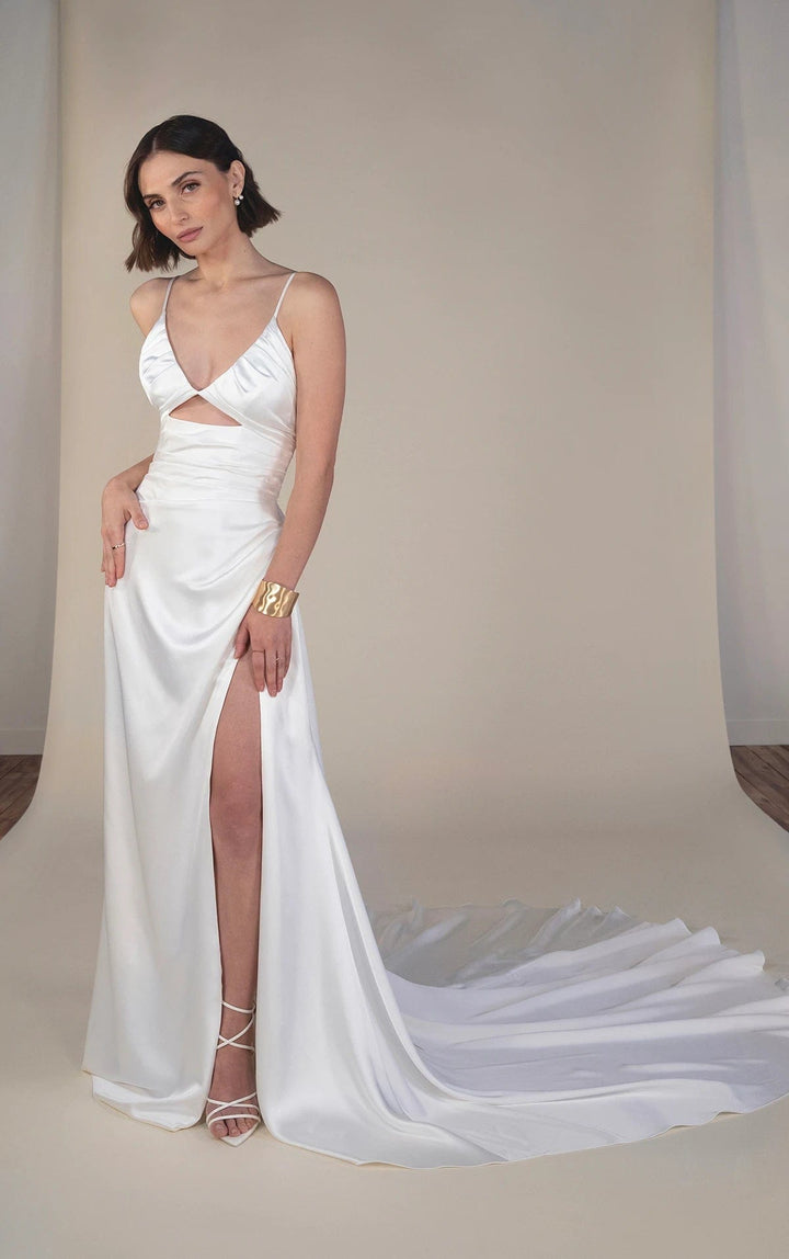 A woman models a white satin formal gown with a thigh-high slit, crossover sweetheart neckline, and a long flowing train against a neutral beige studio backdrop.