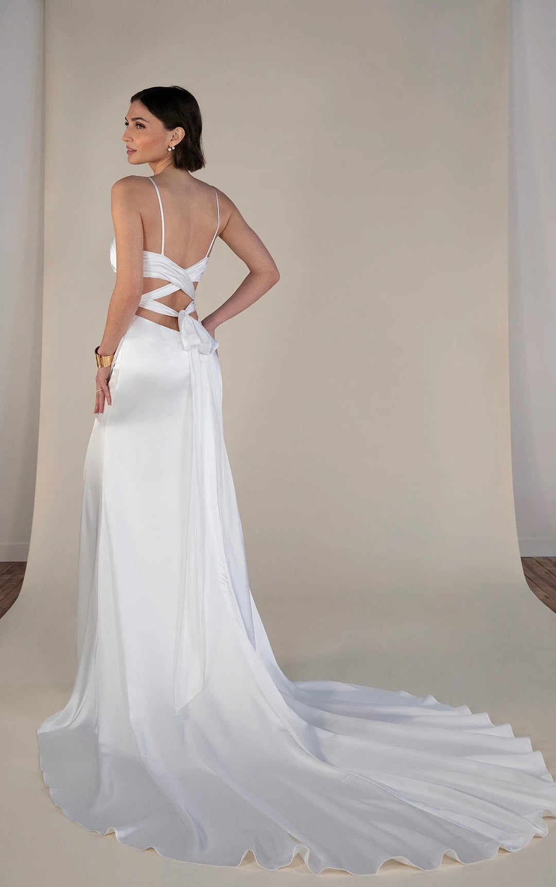 A woman modeling a white satin evening gown from the back, featuring a strappy open back, a large bow at the waist, and a long flowing train against a neutral studio backdrop.