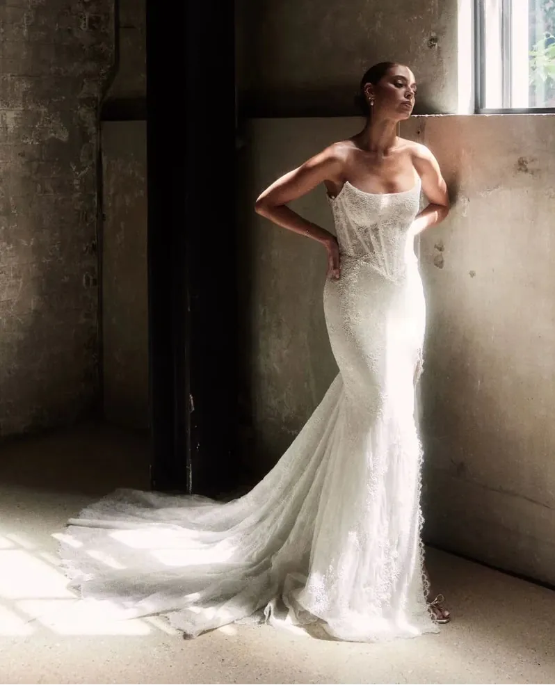 Woman in a strapless wedding dress standing in a sunlit room with concrete walls.