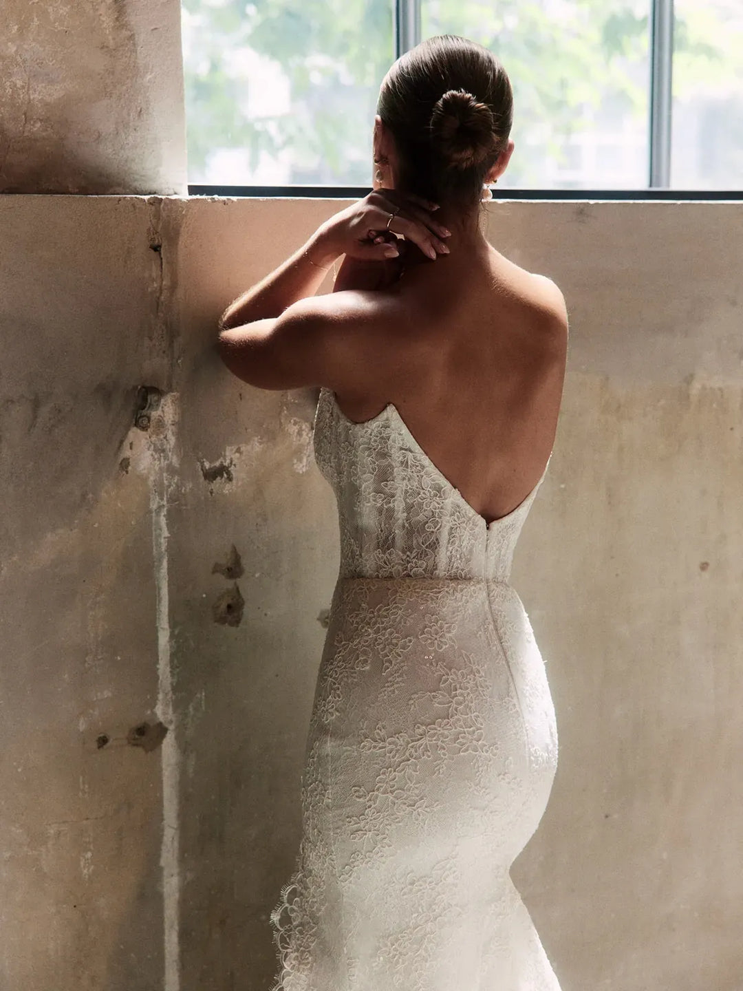 Woman in a lace wedding dress standing against a textured wall with a window in the background
