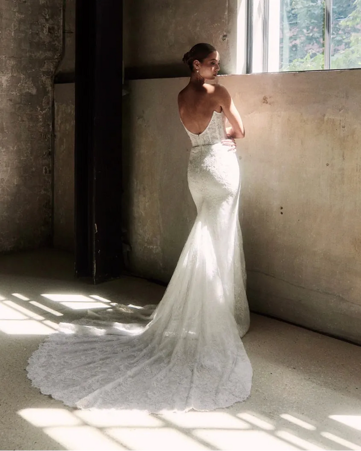 Woman in a white wedding dress standing in a sunlit room with concrete walls.