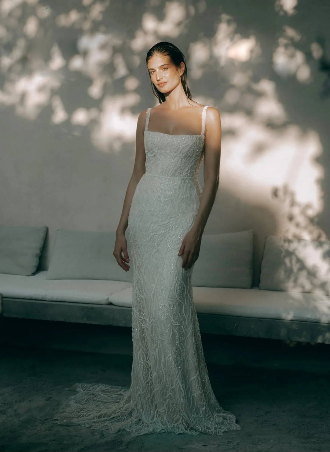 A woman wearing a sleeveless, floor-length white lace gown stands indoors in front of a light-colored wall with dappled sunlight and shadows. A bench is visible behind her.