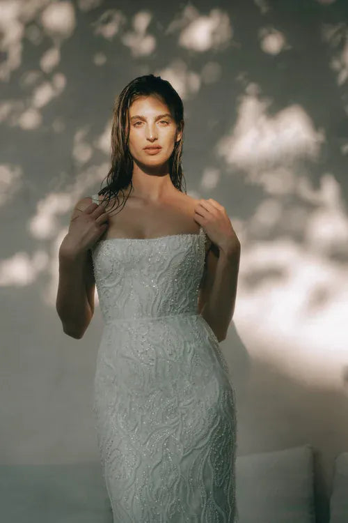 A woman wearing a strapless fitted dress with textured floral lace and subtle beaded embellishments, standing against a softly lit wall with dappled sunlight and shadows.