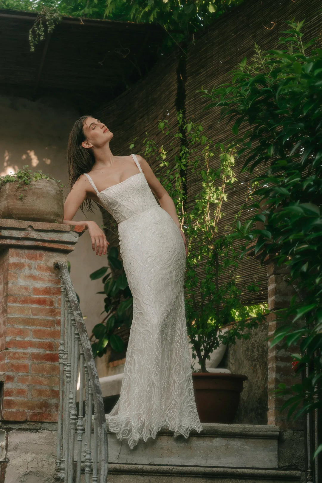 A woman wearing a fitted white lace wedding dress with thin straps and a flared lace hem, standing on outdoor stone steps beside a brick wall and green plants.