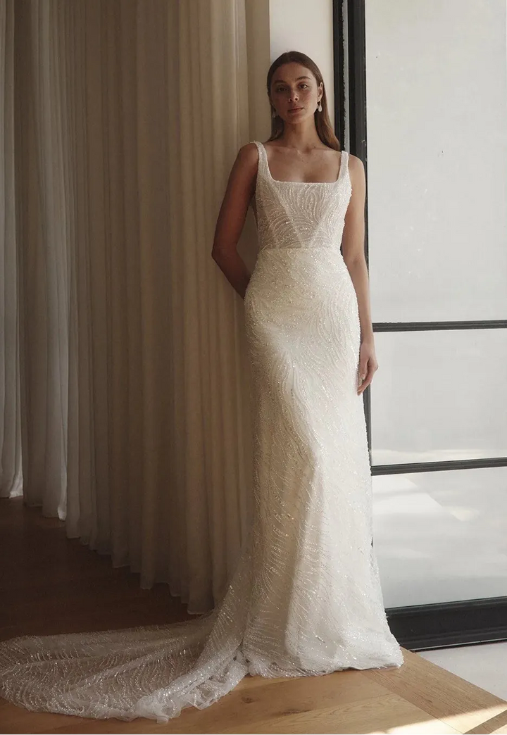 Woman in a white lace wedding dress standing in a room with large windows.