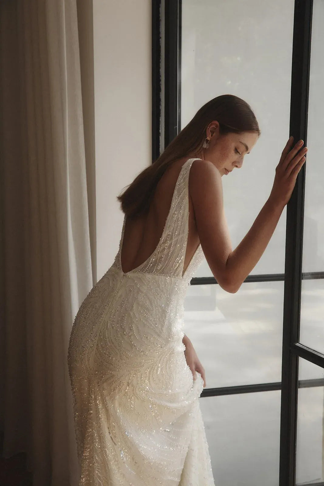 Woman in a white lace wedding dress standing by a window.