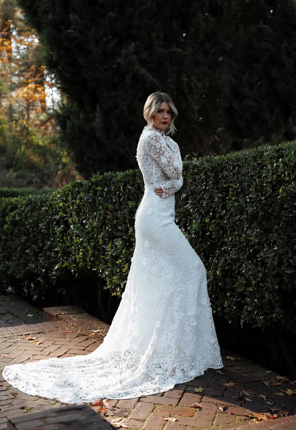 Woman in a white lace wedding dress standing outdoors with greenery in the background