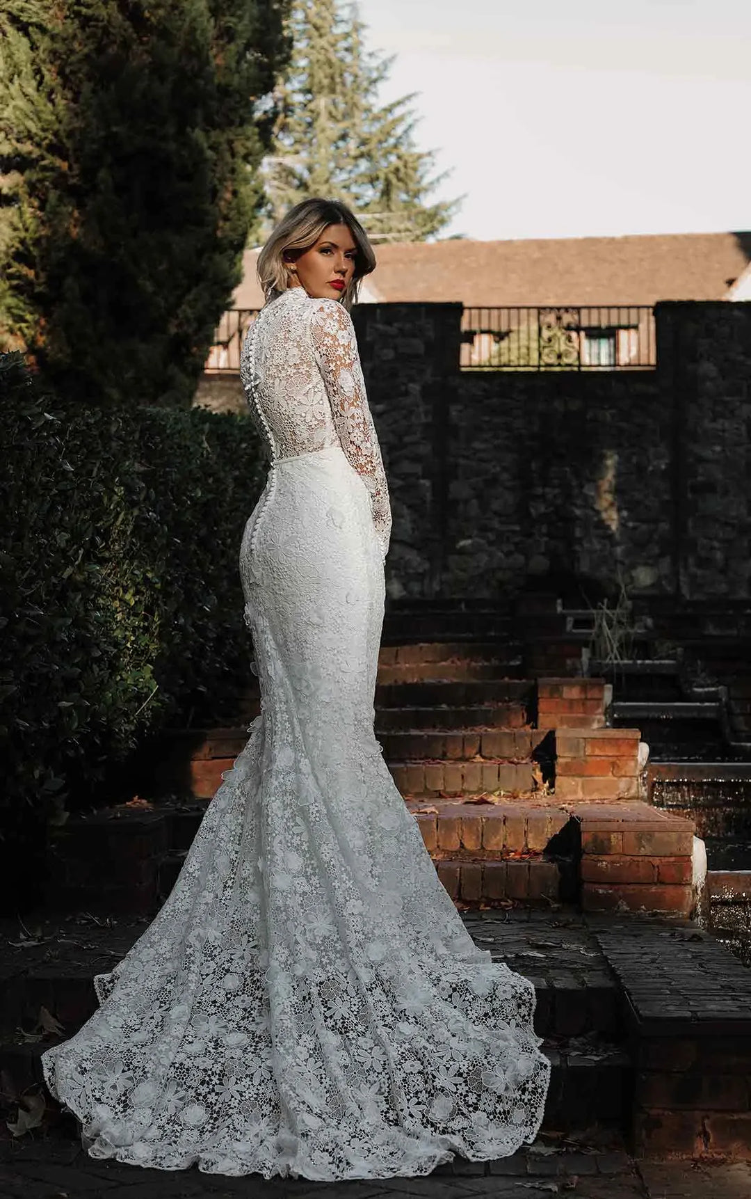 A woman wearing a fitted white lace wedding gown with long sleeves and a dramatic train, standing outdoors on stone steps in front of dark green hedges and a building.
