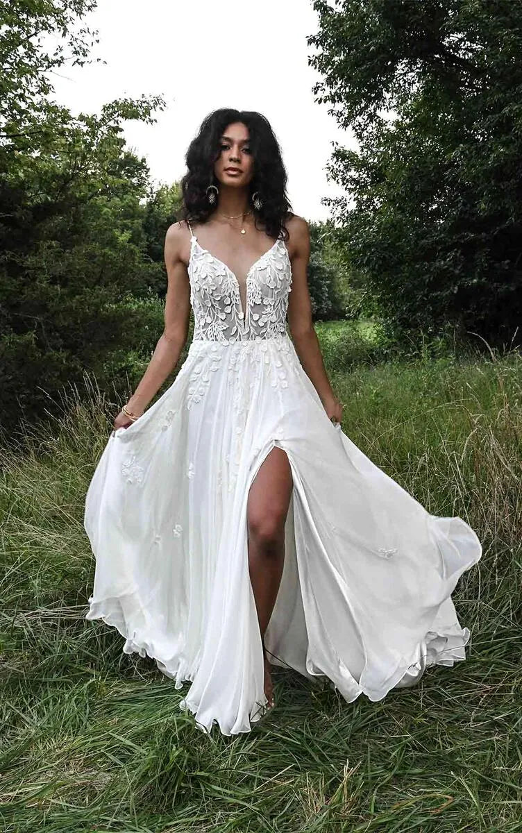 A woman wearing a flowing white wedding dress stands outdoors in tall grass with trees behind her. The dress has a deep V-neckline, delicate lace and bead embellishments on the bodice, thin straps, and a high slit.