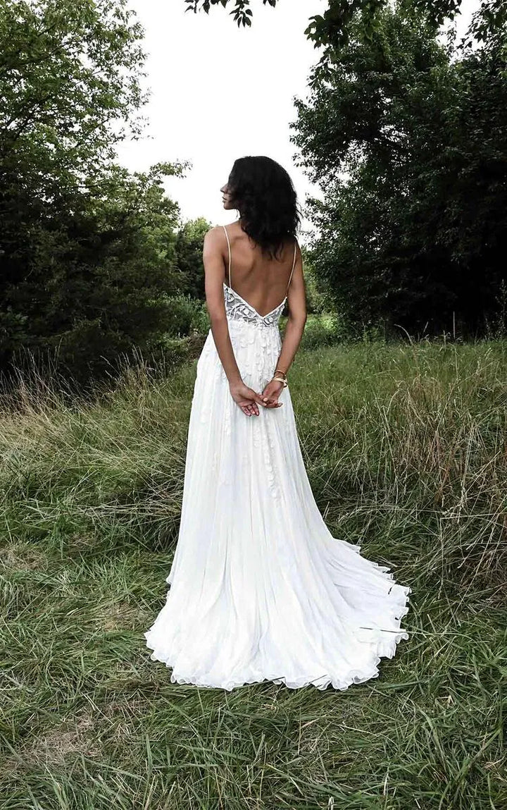 A woman stands outdoors in a grassy field wearing a white wedding dress with a low open back and a long train, photographed from behind.