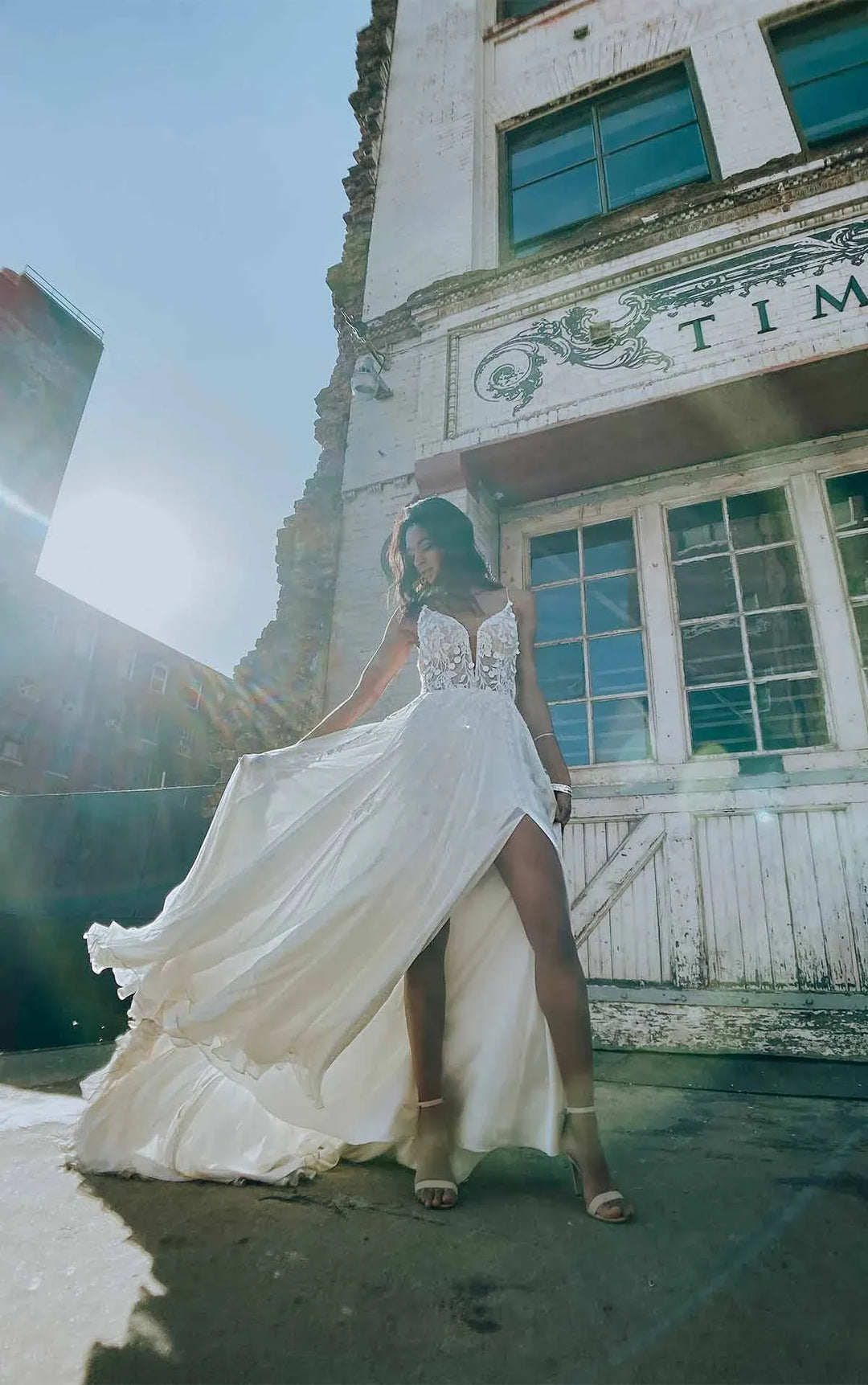 A woman wearing a white sleeveless wedding dress with a high slit and flowing train stands outdoors in front of a weathered building, holding out the skirt to show its volume.