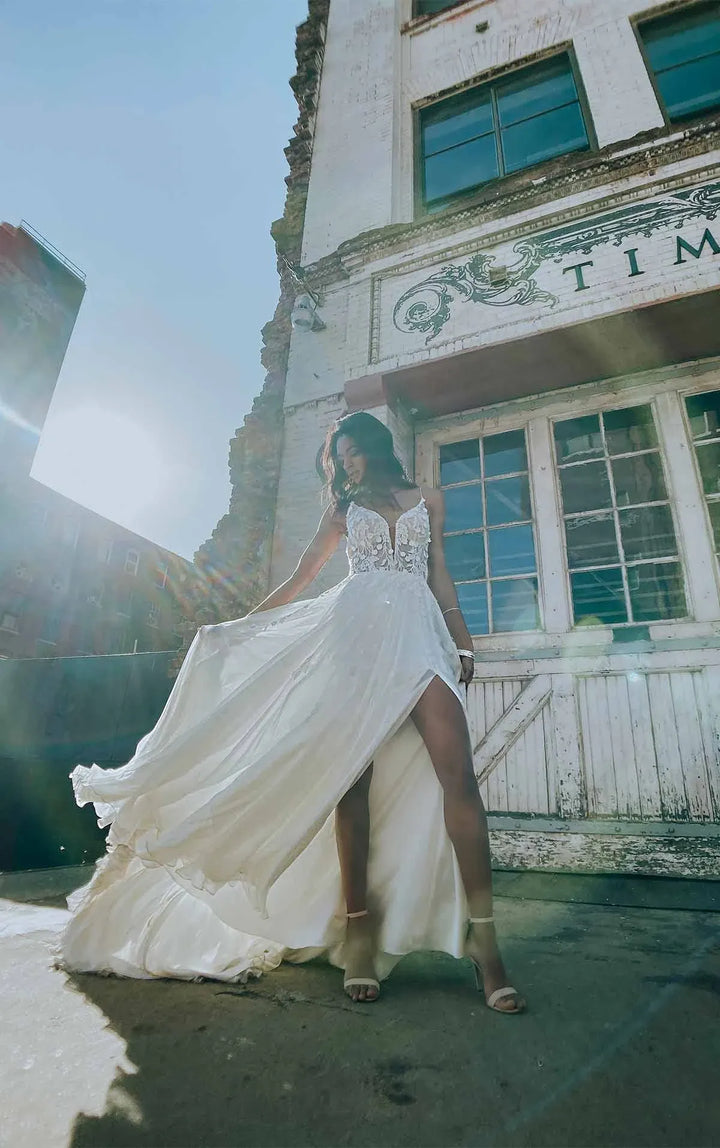 A woman wearing a white sleeveless wedding dress with a high slit and flowing train stands outdoors in front of a weathered building, holding out the skirt to show its volume.