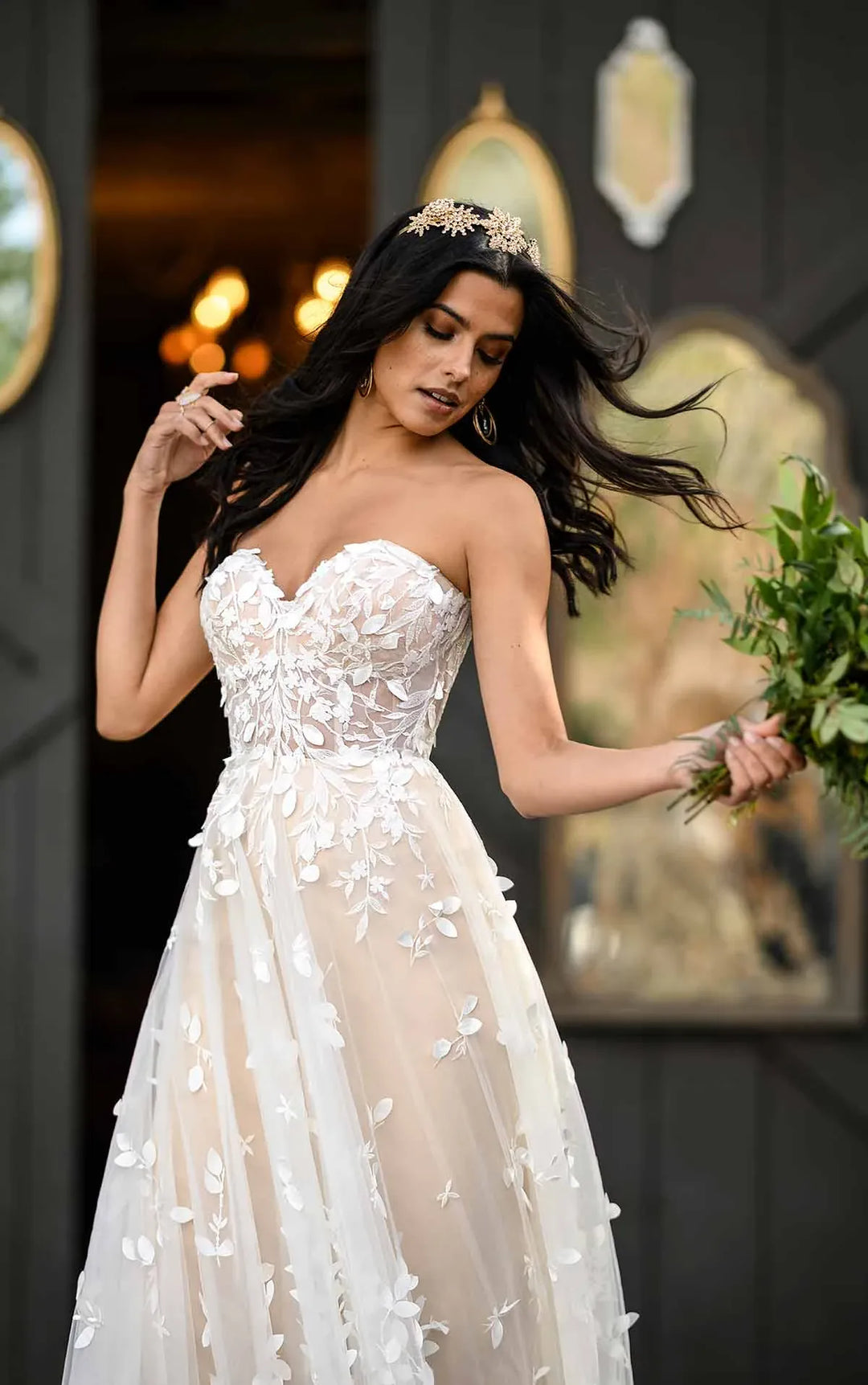 Woman in a white strapless gown with floral details, holding greenery against a dark background.