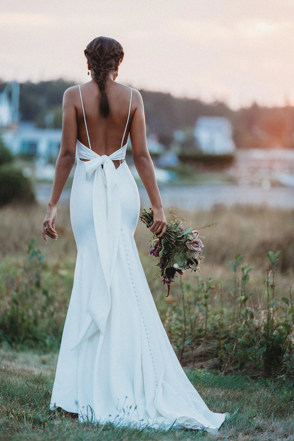 Woman in a white wedding dress with a open back, standing in a field with a sunset in the background.