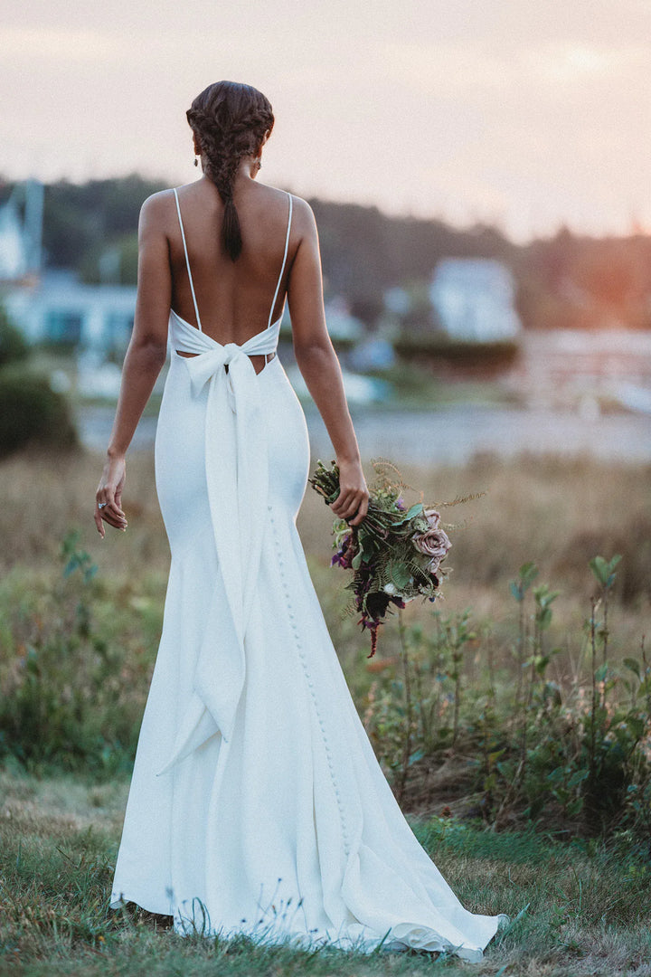 Woman in a white wedding dress with a open back, standing in a field with a sunset in the background.