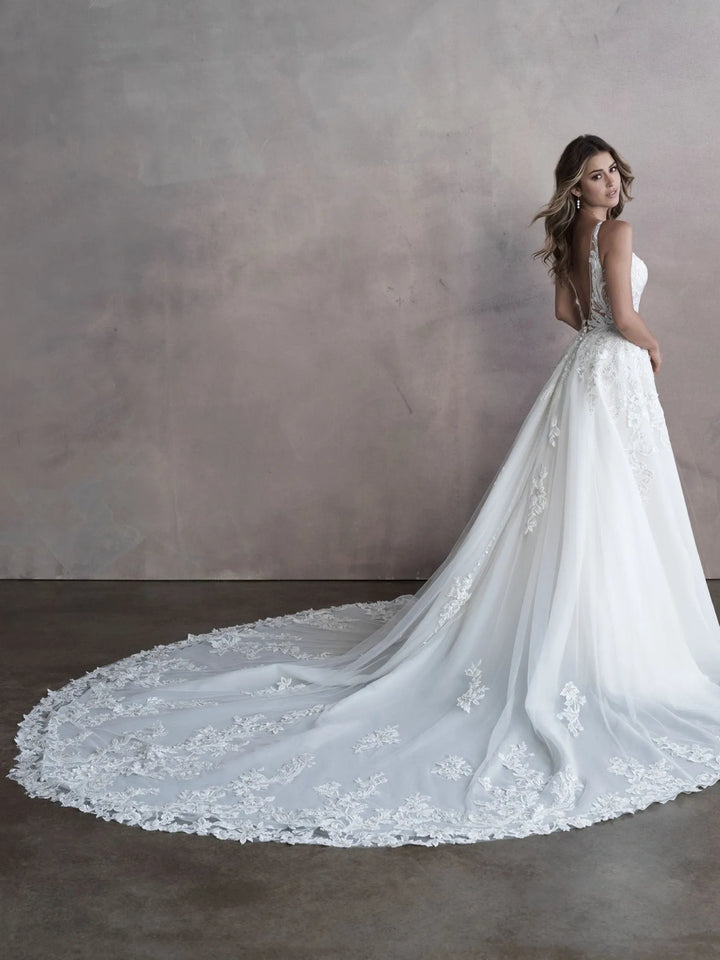 A woman modeling a white sleeveless wedding gown with a long train and floral lace applique, posed against a textured gray studio wall.
