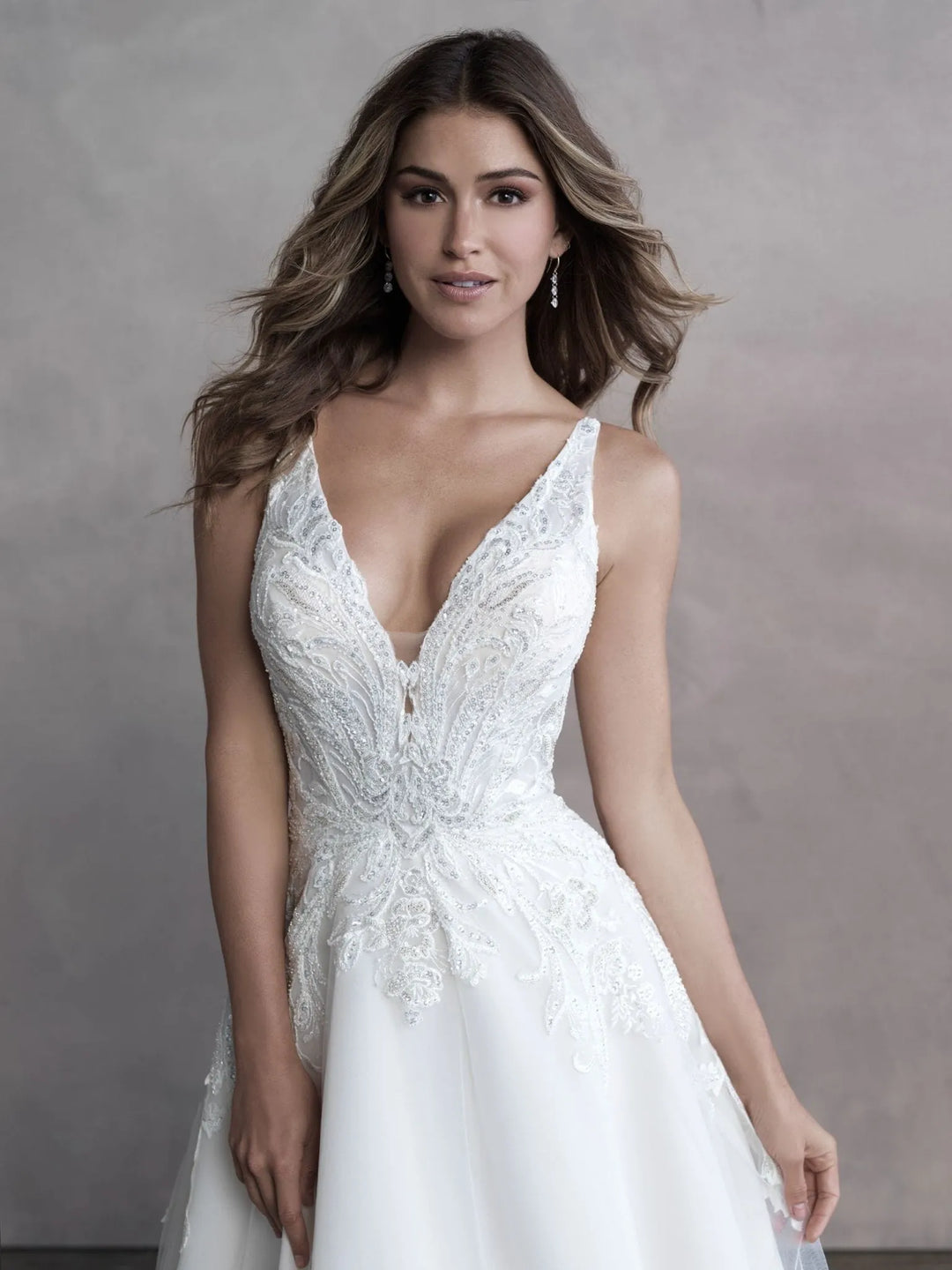A woman modeling a white sleeveless wedding dress with a deep V-neckline, lace bodice, and flowing skirt against a textured gray studio backdrop.
