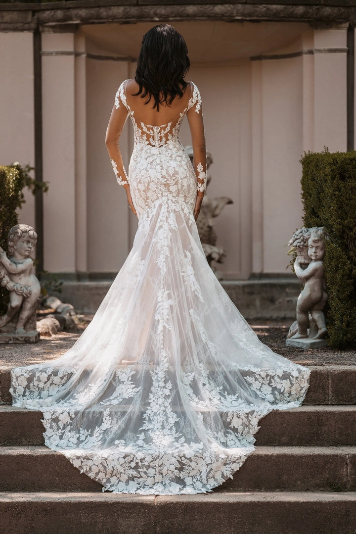Woman in a white lace wedding dress standing on steps with classical architecture and statues in the background.