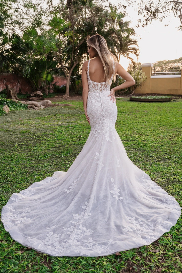 A woman wearing a white lace mermaid-style wedding gown is photographed from behind outdoors on grass, with trees and a building visible in the background.