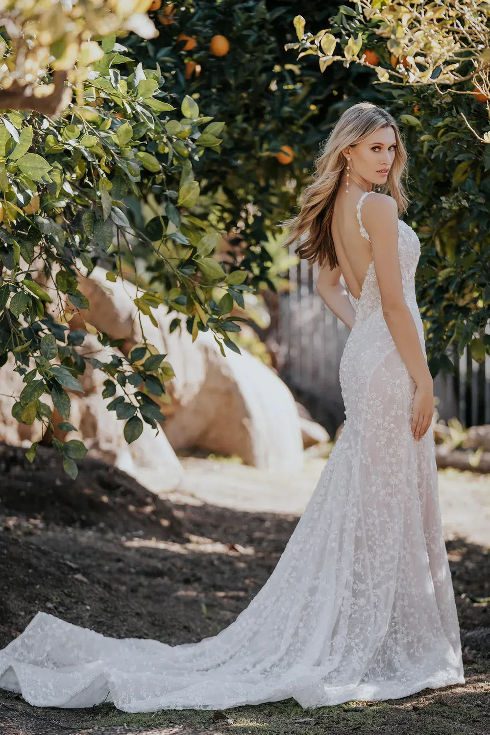 Woman in a white lace wedding dress standing in a garden with trees and rocks in the background.