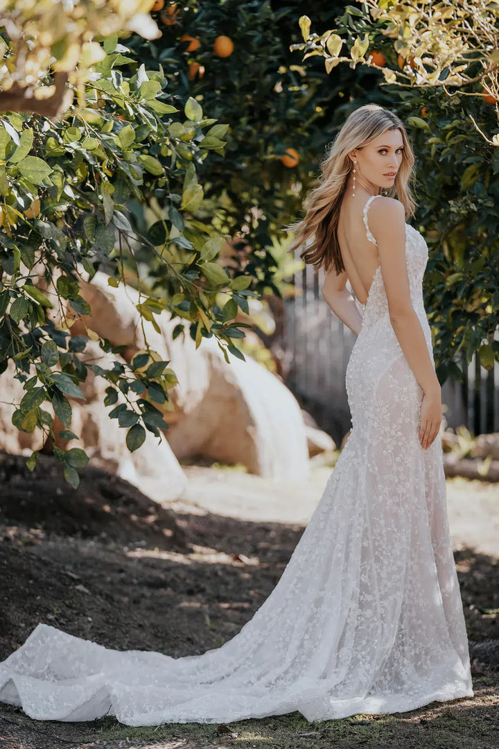 Woman in a white lace wedding dress standing in a garden with trees and rocks in the background.