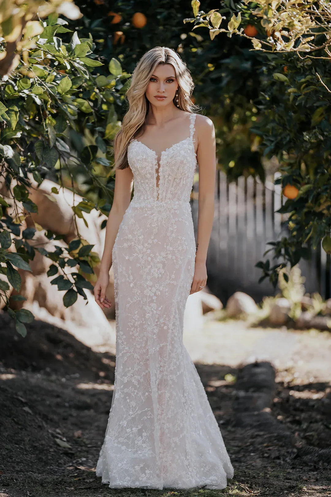 A woman wearing a fitted white lace mermaid-style wedding gown stands outdoors in an orchard with leafy trees and sunlight filtering through.