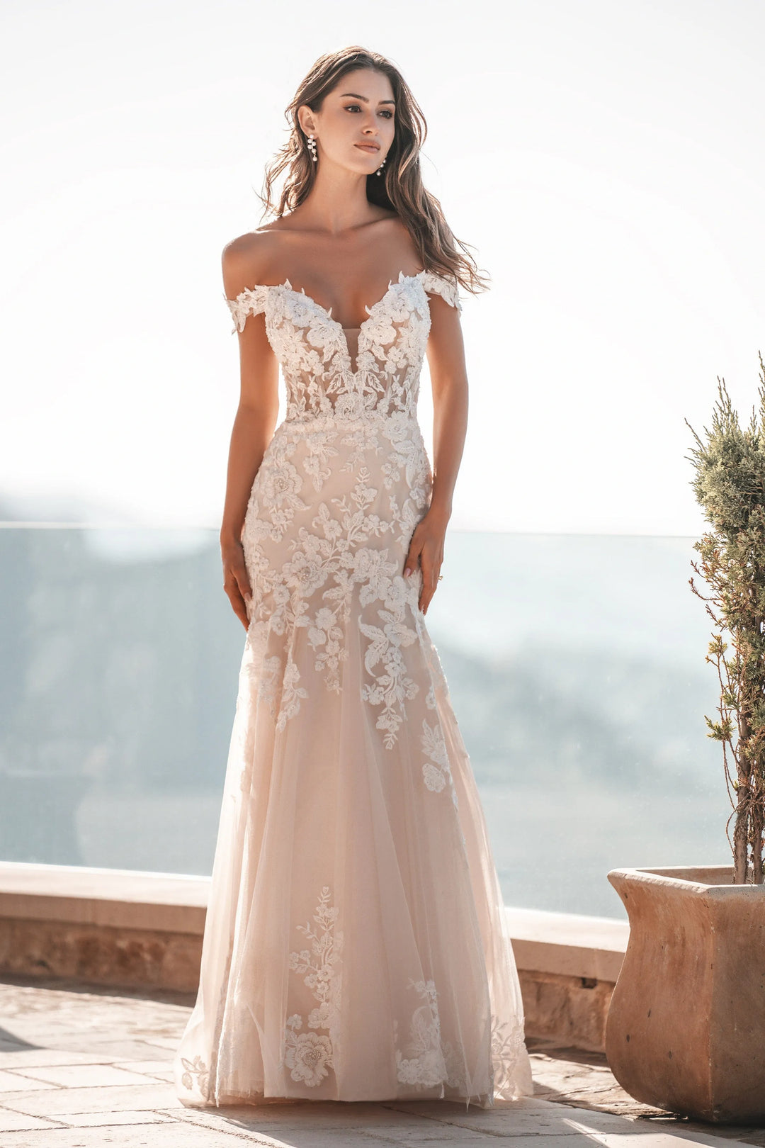 A woman modeling an off-the-shoulder mermaid-style wedding gown with floral lace applique and a sheer tulle skirt, photographed outdoors on a sunlit terrace with a blurred scenic background.
