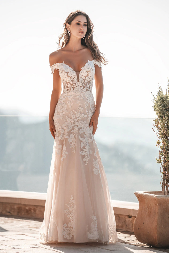 A woman modeling an off-the-shoulder mermaid-style wedding gown with floral lace applique and a sheer tulle skirt, photographed outdoors on a sunlit terrace with a blurred scenic background.
