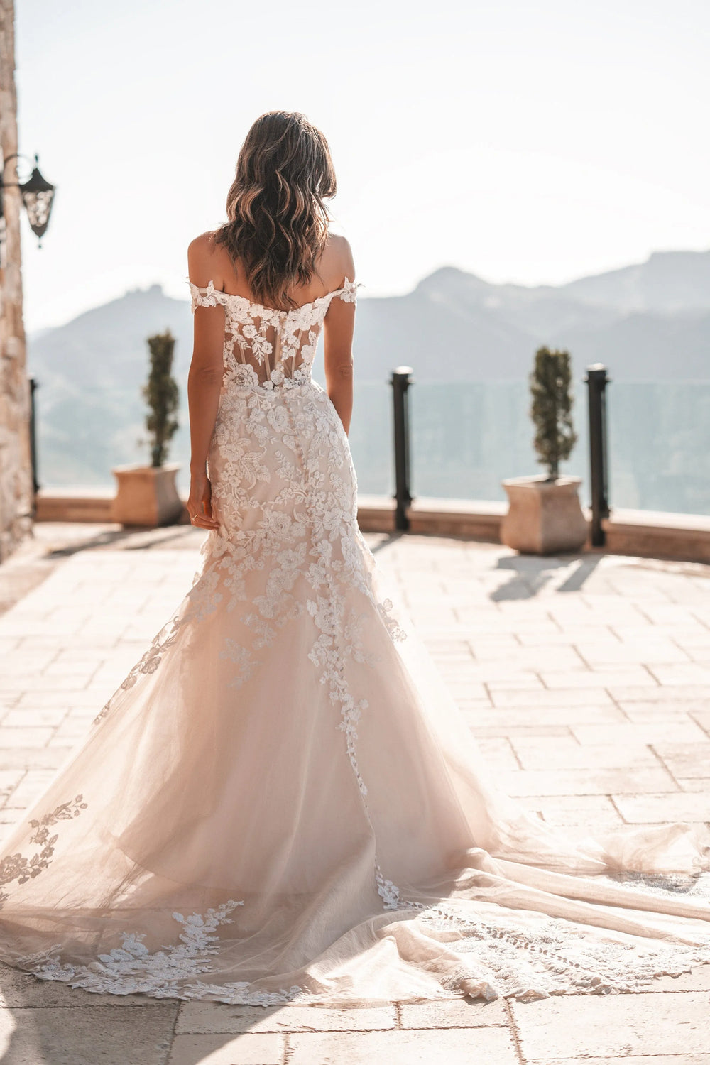 A woman stands on a sunlit terrace wearing a bridal gown from behind, with mountains and a railing in the background. The dress is off-the-shoulder, fitted, and features a long train with floral lace detailing.