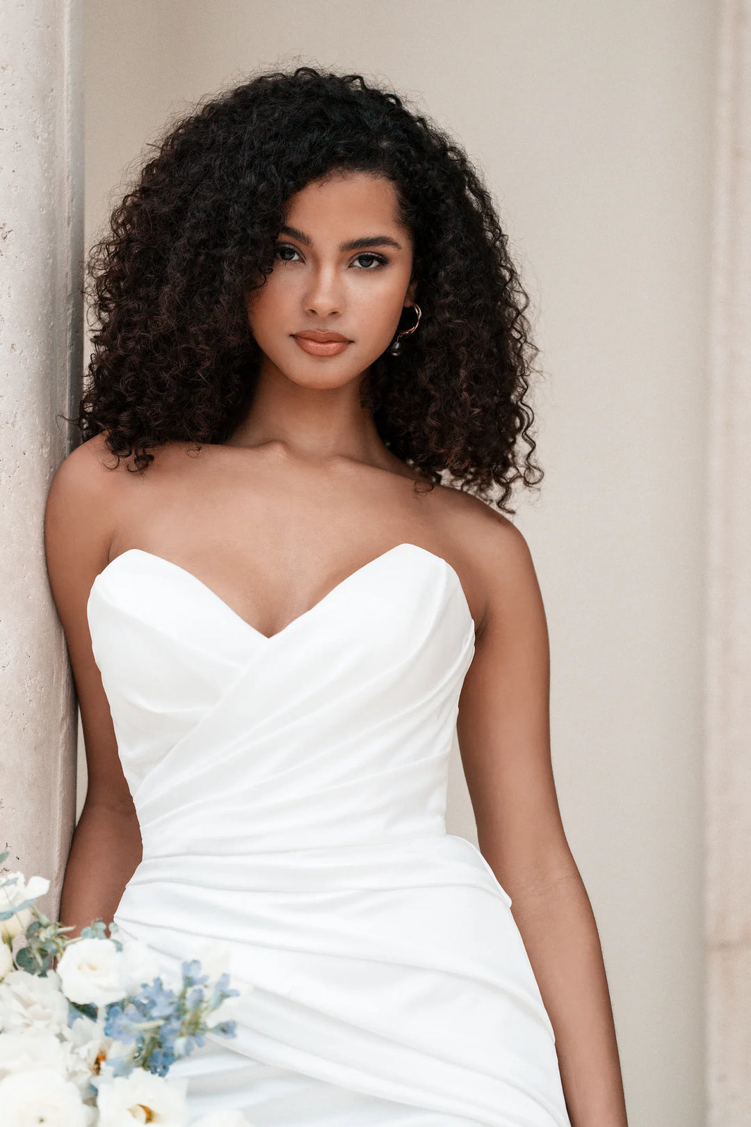 A woman with curly black hair wearing a strapless white sweetheart-neckline wedding dress, holding a bouquet of white and light blue flowers, posed against a neutral beige wall.