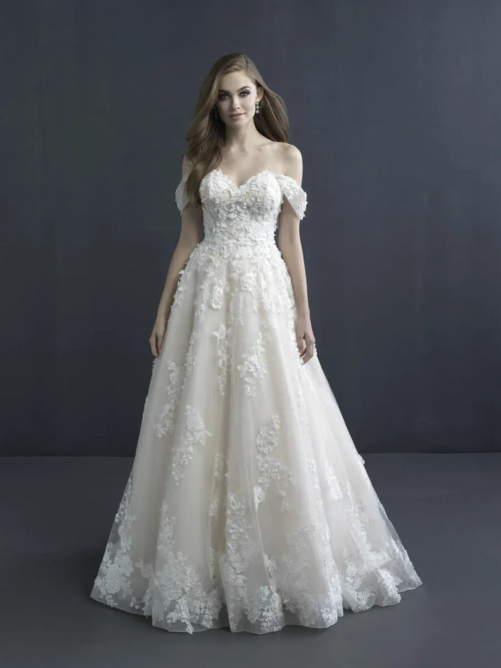 A woman modeling an off-the-shoulder A-line wedding gown with a sweetheart neckline, fitted lace bodice, and voluminous tulle skirt with floral lace applique, photographed against a dark gray studio backdrop.