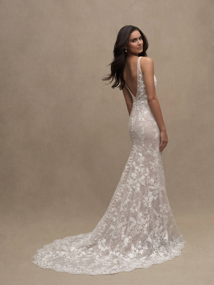 A woman modeling a white lace mermaid-style wedding gown with a deep V back and long train, photographed against a plain warm beige studio backdrop.