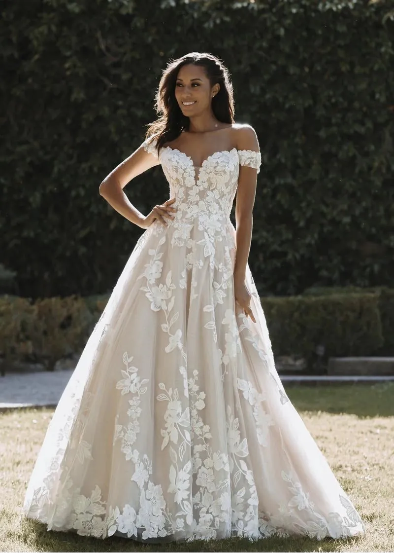 A woman wearing a voluminous off-the-shoulder A-line wedding gown with 3D floral applique, standing outdoors on grass with a green hedge in the background.