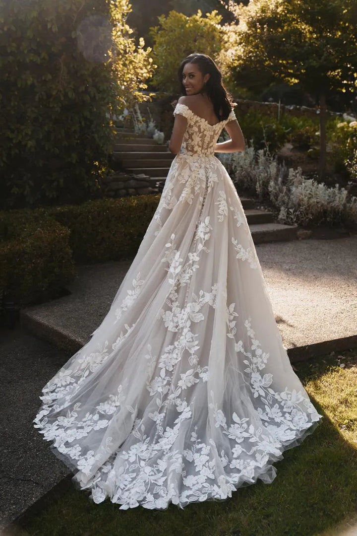 A woman wearing a voluminous off-white wedding gown with floral lace applique and cap sleeves, standing outdoors on a garden path with trees and stone steps in the background.