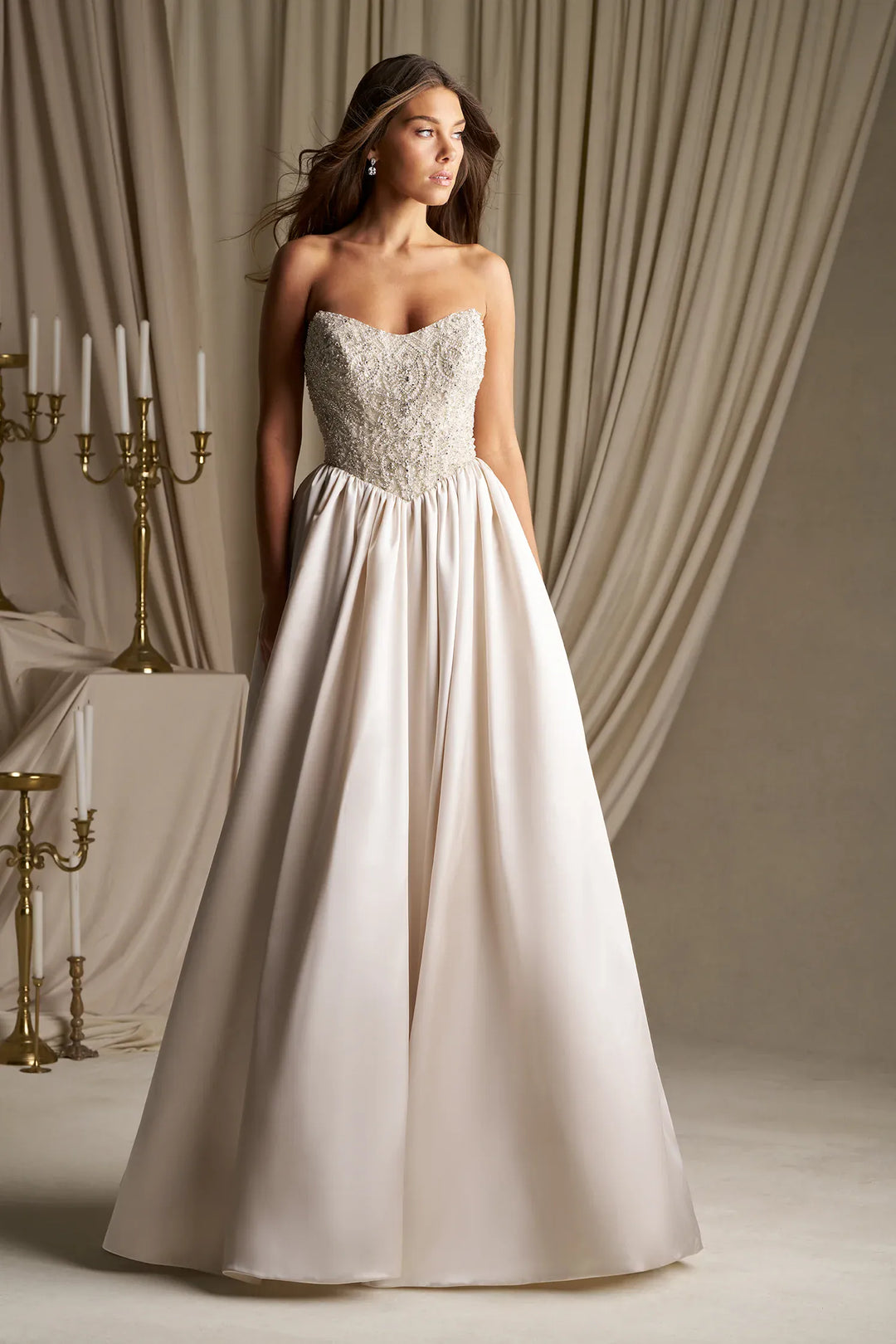 A woman modeling a strapless A-line wedding gown with a beaded sweetheart bodice and a full flowing skirt, posed in front of draped beige curtains and gold candelabras.