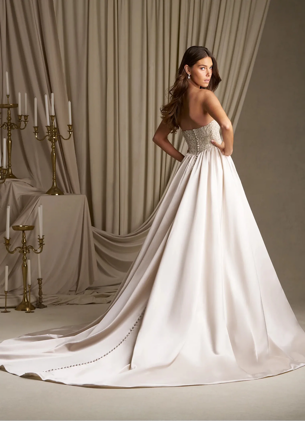 A woman modeling a strapless white wedding gown with a fitted embellished bodice and a long flowing train, posed in front of draped beige curtains and gold candelabras.