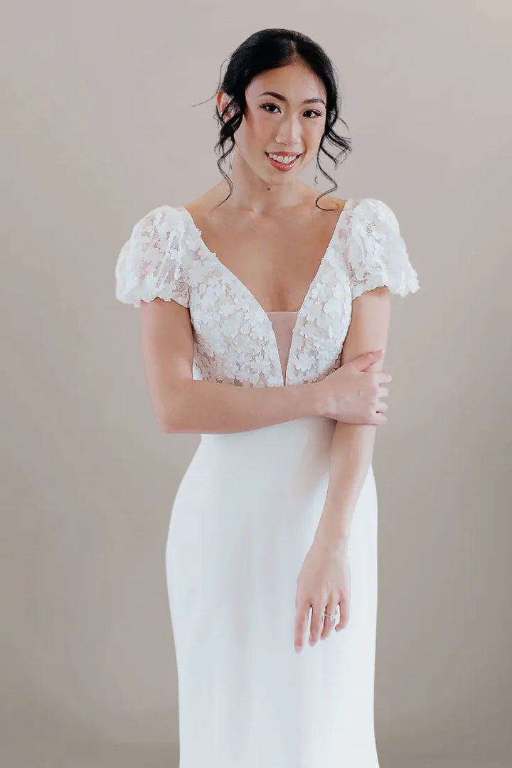 A woman modeling a white wedding dress with a lace bodice, short puff sleeves, and a smooth satin skirt, posed against a neutral beige studio background.