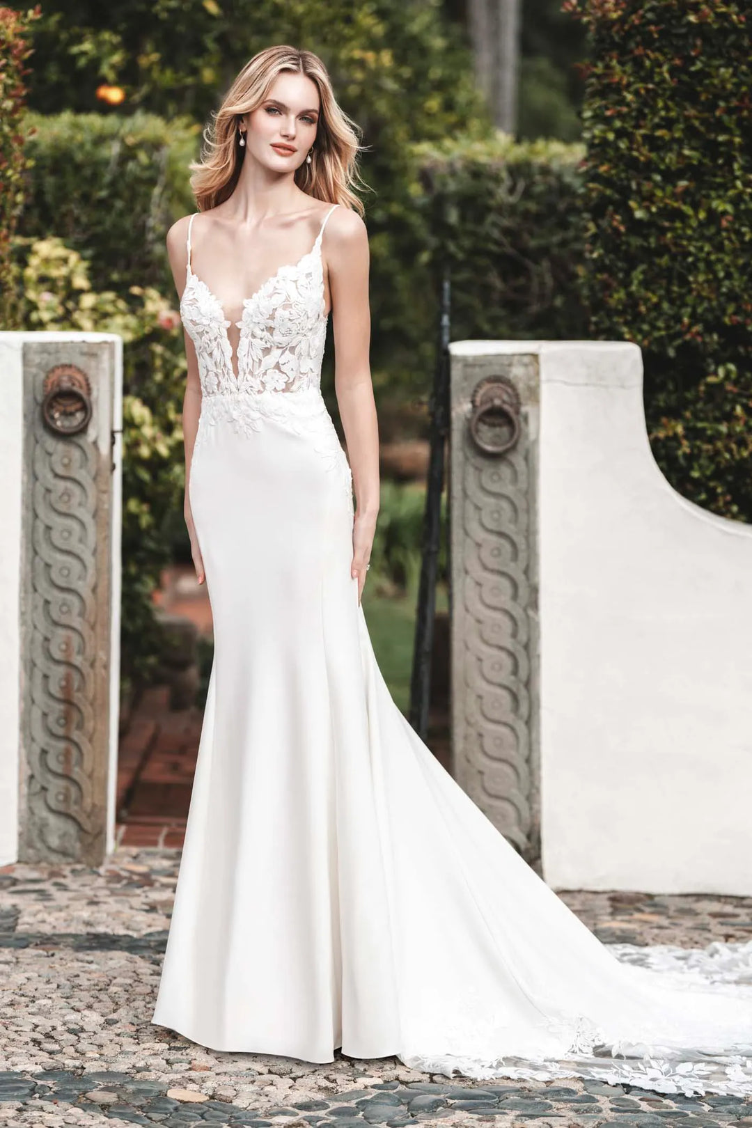 A woman modeling a white mermaid-style wedding gown with thin straps, a lace-embellished bodice, and a long train in an outdoor garden setting.
