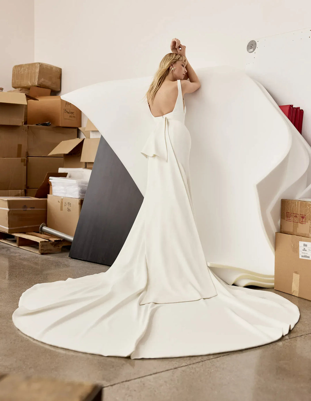 Woman in a white dress standing in an industrial setting with boxes and a large mirror.