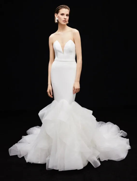 A model wearing a strapless white mermaid-style wedding gown with a deep V neckline, fitted bodice, and a voluminous layered tulle skirt, posed against a solid black background.