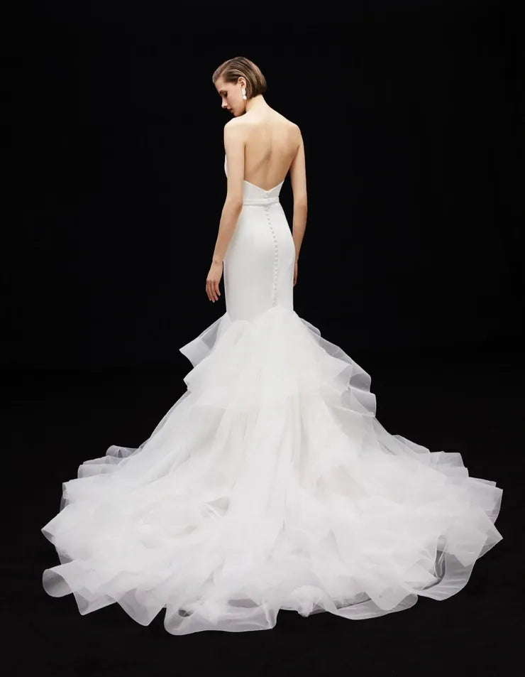 A model shown from the back wearing a white strapless mermaid-style wedding gown with a fitted bodice, open back, and a very full layered tulle skirt, photographed against a black studio background.