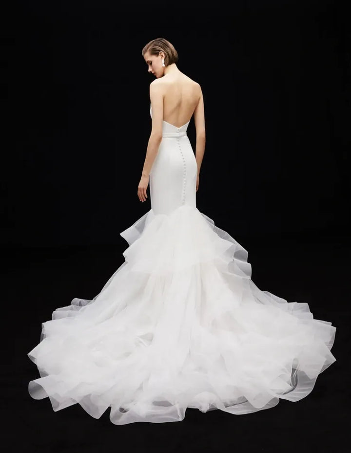 A model shown from the back wearing a white strapless mermaid-style wedding gown with a fitted bodice, open back, and a very full layered tulle skirt, photographed against a black studio background.