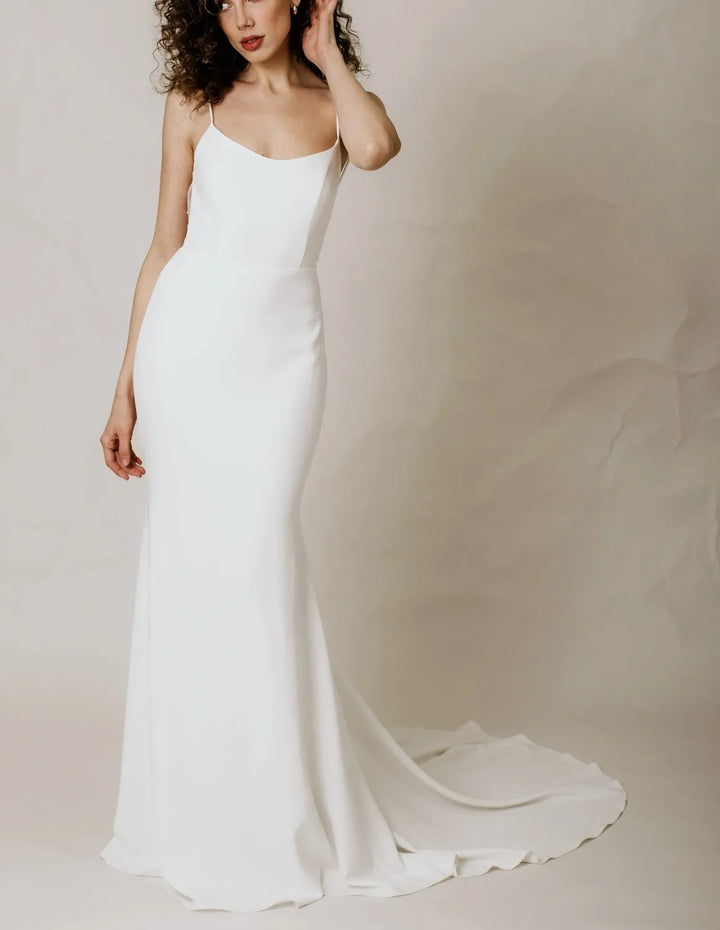 A woman modeling a floor-length white wedding gown with thin spaghetti straps, a fitted silhouette, and a slight train against a soft beige studio backdrop.