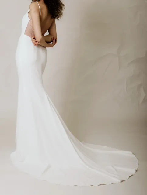 A woman modeling a white sleeveless wedding dress with a sheer nude back panel and a long flowing train, photographed against a soft beige studio backdrop.