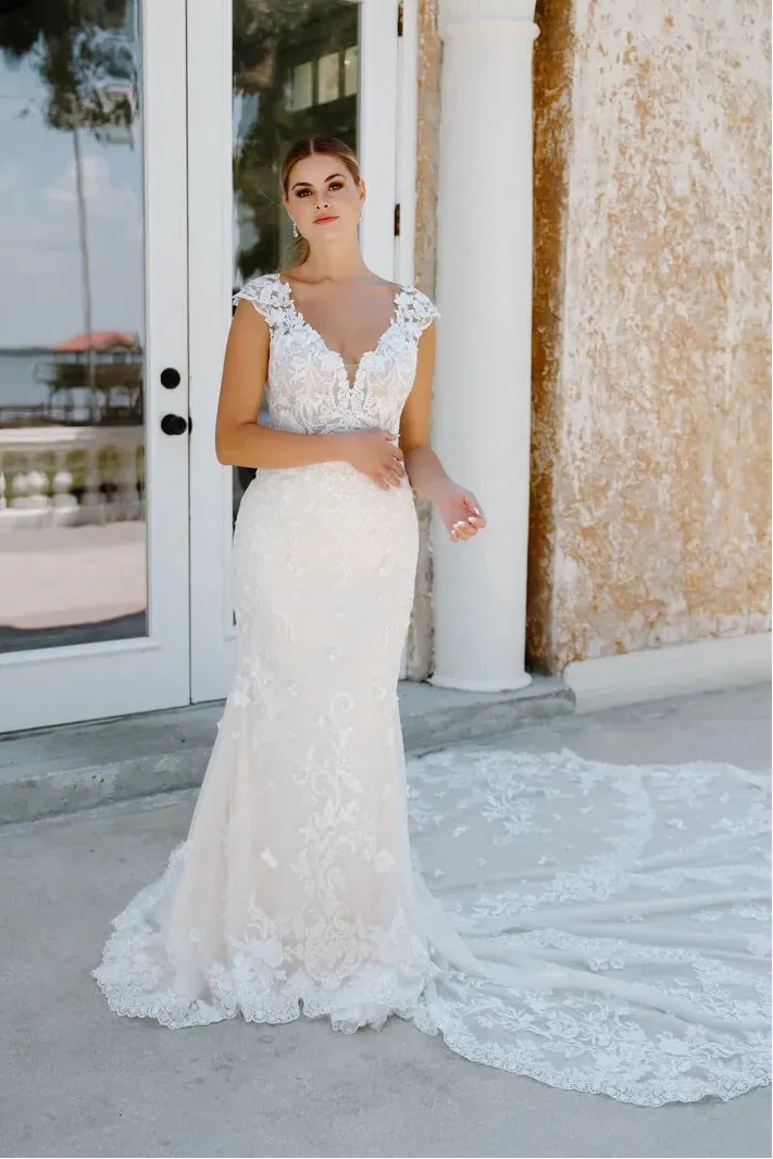 A woman wearing a sleeveless lace mermaid-style wedding gown with a deep V-neckline and cap sleeves, standing outdoors in front of white double doors and a textured beige wall. The dress has a fitted bodice, flared lace skirt, and a long matching train on the ground.