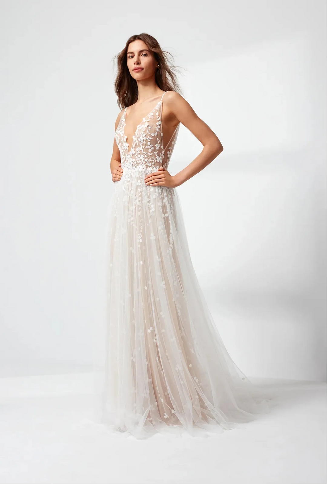 A woman modeling a sleeveless, floor-length white tulle wedding gown with intricate bead and sequin embellishments, set against a clean white studio background.