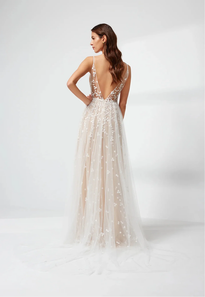 A woman modeling a sleeveless, backless A-line wedding gown from the back, featuring a deep V-neckline, thin straps, and a voluminous tulle skirt adorned with intricate beadwork and sequins, set against a clean white studio background.